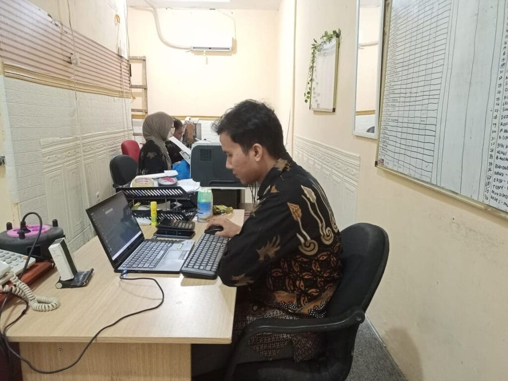 Seorang karyawan PT Solusi Mandiri Rekananda, perusahaan jasa konsultan pajak di Bekasi, sedang bekerja di meja dengan fokus, dikelilingi oleh peralatan kantor dan papan informasi di dinding.